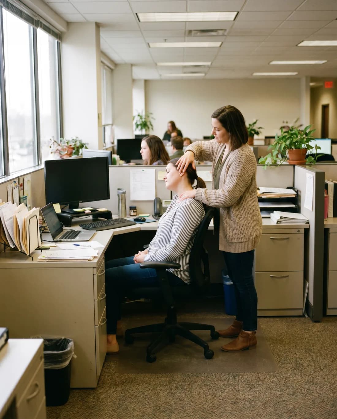 A facilitator guiding an office worker through a gentle desk-side stretch by bright office windows. Illustrative.